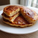 A stack of three golden-brown banana pancakes drizzled with syrup, one cut open to show the fluffy interior.