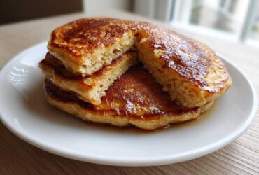 A stack of three golden-brown banana pancakes drizzled with syrup, one cut open to show the fluffy interior.