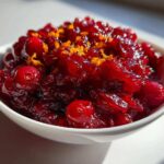 Close-up of vibrant, glistening homemade cranberry sauce topped with bright orange zest in a white bowl.