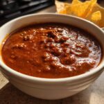 A close-up of thick, rich, reddish-brown chili cheese dip served in a white bowl with tortilla chips visible in the background.