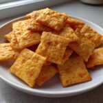 A white bowl filled with crispy, square, orange homemade cheese crackers, ready for snacking.