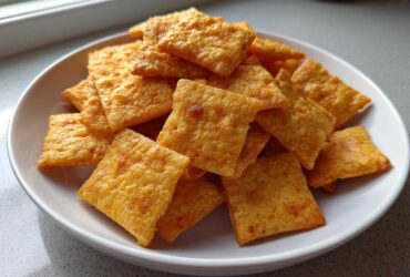A white bowl filled with crispy, square, orange homemade cheese crackers, ready for snacking.