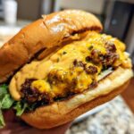 Close-up of a hand holding a juicy smash burger generously topped with melted cheese and orange burger sauce.