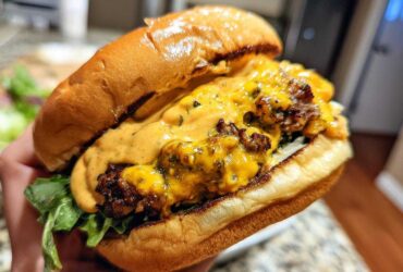 Close-up of a hand holding a juicy smash burger generously topped with melted cheese and orange burger sauce.