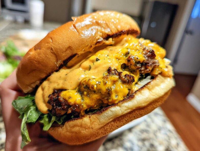 Close-up of a hand holding a juicy smash burger generously topped with melted cheese and orange burger sauce.