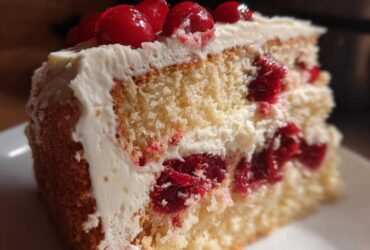 Close-up of a moist slice of cherry cake with white frosting and whole cherries on top.