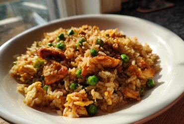 A close-up shot of a white bowl filled with savory chicken fried rice, featuring pieces of chicken and bright green peas.
