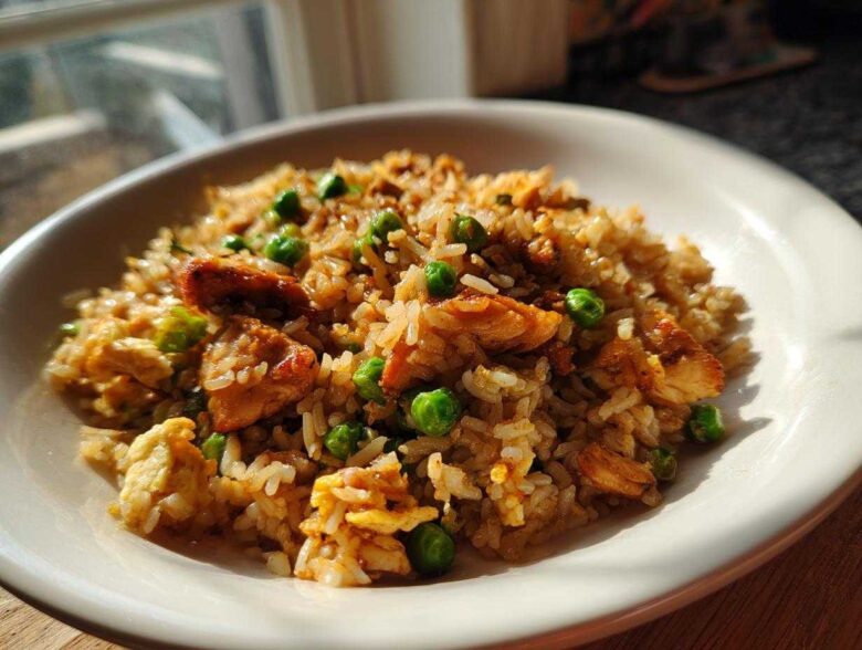 A close-up shot of a white bowl filled with savory chicken fried rice, featuring pieces of chicken and bright green peas.