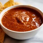 Close-up of rich, thick chili cheese dip in a white bowl, served with tortilla chips nearby.