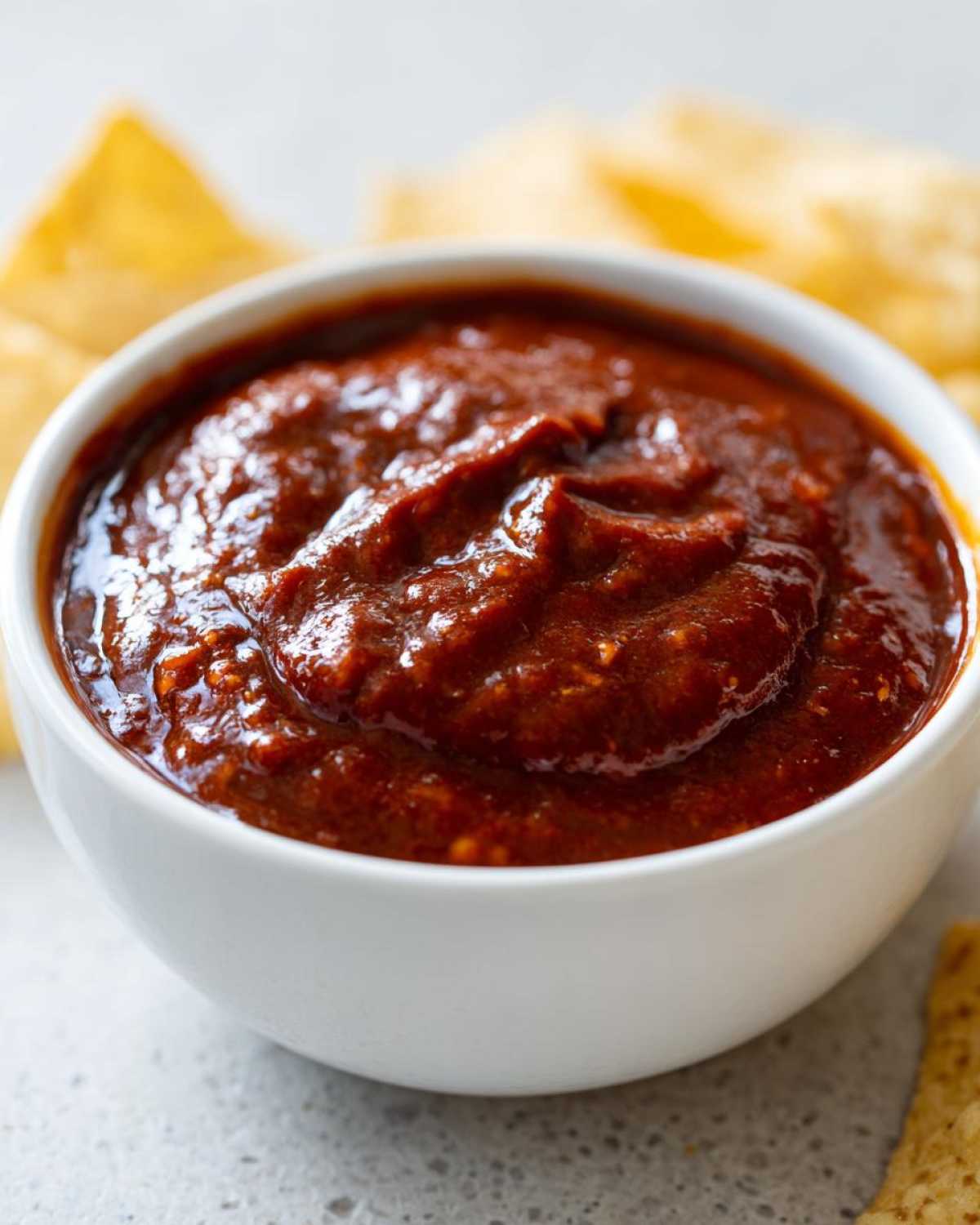 Close-up of a thick, rich red chili cheese dip served in a white bowl with tortilla chips blurred in the background.