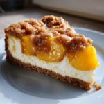 A close-up slice of peach cobbler cheesecake showing a graham cracker crust, creamy filling, and caramelized peaches with a crumble topping.