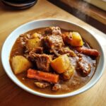 Close-up of a white bowl filled with tender chunks of pork stew, potatoes, and carrots in a rich brown gravy.