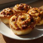 Four freshly baked sausage pinwheels with melted cheese topping arranged on a white plate.