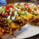 A close-up shot of a slice of amazing taco pizza being held up, showing seasoned ground beef, melted cheddar cheese, lettuce, tomatoes, and sour cream drizzle.