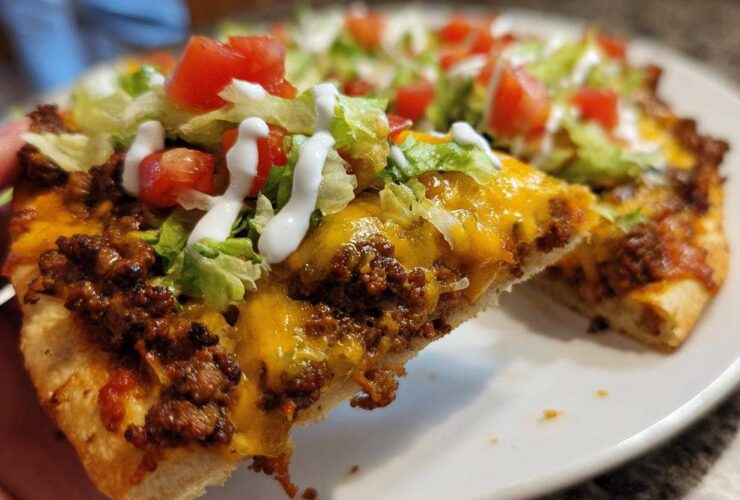 A close-up shot of a slice of amazing taco pizza being held up, showing seasoned ground beef, melted cheddar cheese, lettuce, tomatoes, and sour cream drizzle.