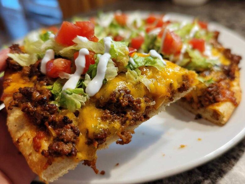 A close-up shot of a slice of amazing taco pizza being held up, showing seasoned ground beef, melted cheddar cheese, lettuce, tomatoes, and sour cream drizzle.