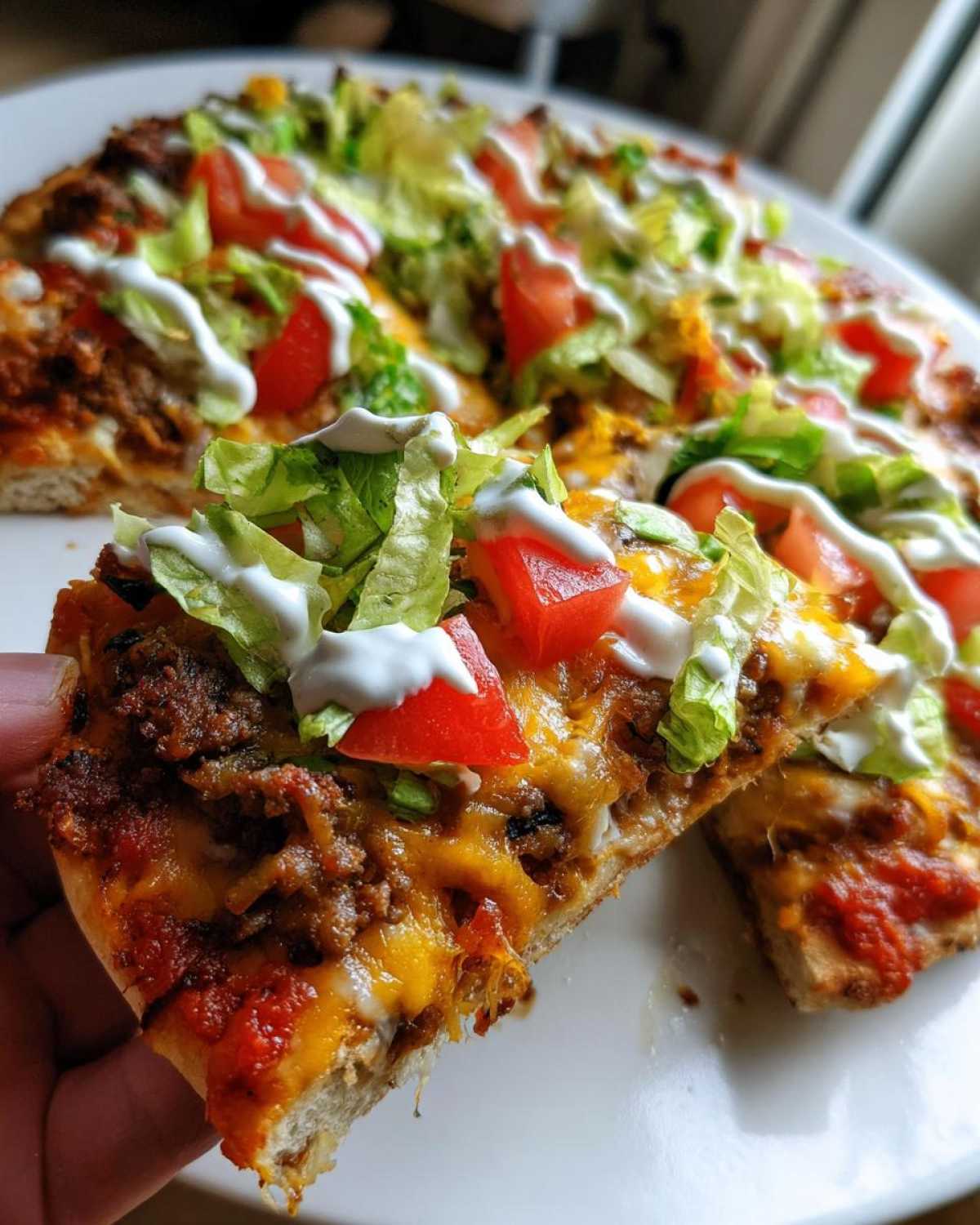 A hand holding a slice of taco pizza topped with seasoned beef, cheese, lettuce, tomato, and sour cream drizzle.