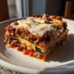 A hearty, layered slice of zucchini lasagna featuring meat sauce and melted mozzarella cheese on a white plate.