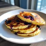 A stack of three golden brown blueberry coconut flour pancakes drizzled with syrup and topped with melting butter.