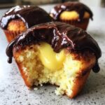 A close-up of a boston cream pie cupcakes with a bite taken out, revealing rich yellow pastry cream filling and dark chocolate ganache topping.