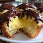 A close-up of a boston cream pie cupcake cut in half, showing rich vanilla cake filled with creamy yellow custard and topped with chocolate ganache.