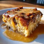 Close-up of a rich slice of bread pudding with bourbon sauce generously drizzled over the top and pooling on the white plate.