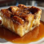 A decadent square of bread pudding with bourbon sauce generously poured over the top and pooling on the white plate.