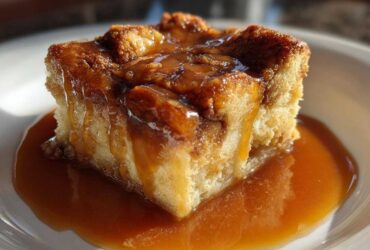 A decadent square of bread pudding with bourbon sauce generously poured over the top and pooling on the white plate.