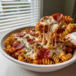 A spoonful of cheesy pizza pasta bake is lifted from a white bowl, showing a dramatic cheese pull.