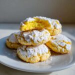 A stack of chewy lemon cookies heavily dusted with powdered sugar, one cookie broken open to show the bright yellow interior.
