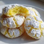 A stack of chewy lemon cookies covered in cracked powdered sugar, showing a soft yellow interior.