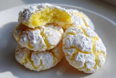 A stack of chewy lemon cookies covered in cracked powdered sugar, showing a soft yellow interior.