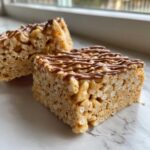 Two chewy peanut butter rice krispie treats squares, drizzled with melted chocolate, sitting on a marble surface.