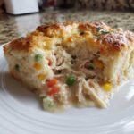 A close-up of a serving of chicken pot pie casserole showing shredded chicken, vegetables, and creamy sauce under a golden biscuit topping.