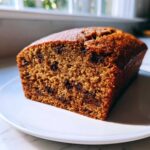 A close-up view of a moist slice of vegan banana bread studded with dark chocolate chips, resting on a white plate.