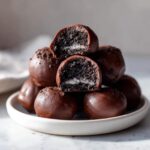 A stack of rich, chocolate-coated Oreo balls, with two cut in half showing the dark cookie center and white filling.