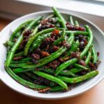 A close-up of vibrant green beans tossed in a dark, savory glaze and topped with crispy bacon bits.