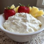 A white bowl filled with creamy fruit dip, garnished with fresh strawberries and pineapple slices.