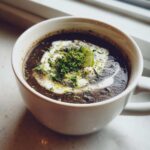 A steaming white mug filled with creamy black bean soup, topped with a swirl of cream, fresh parsley, and a lime wedge.