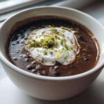 Close-up of creamy black bean soup in a white mug, topped with sour cream and bright green lime zest.