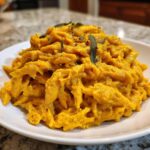 A plate of penne pasta coated thickly in bright orange butternut squash pasta sauce, topped with fresh sage.