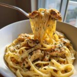 A fork lifting strands of creamy chicken pasta with chunks of seasoned chicken from a white bowl.