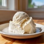 A close-up of two scoops of creamy, white cottage cheese ice cream melting slightly on a light plate.