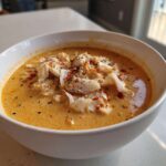A white bowl filled with creamy, orange-hued crab soup, topped generously with white crab meat and red pepper flakes.