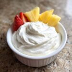 A white bowl filled with creamy fruit dip, garnished with fresh strawberry slices and pineapple chunks.