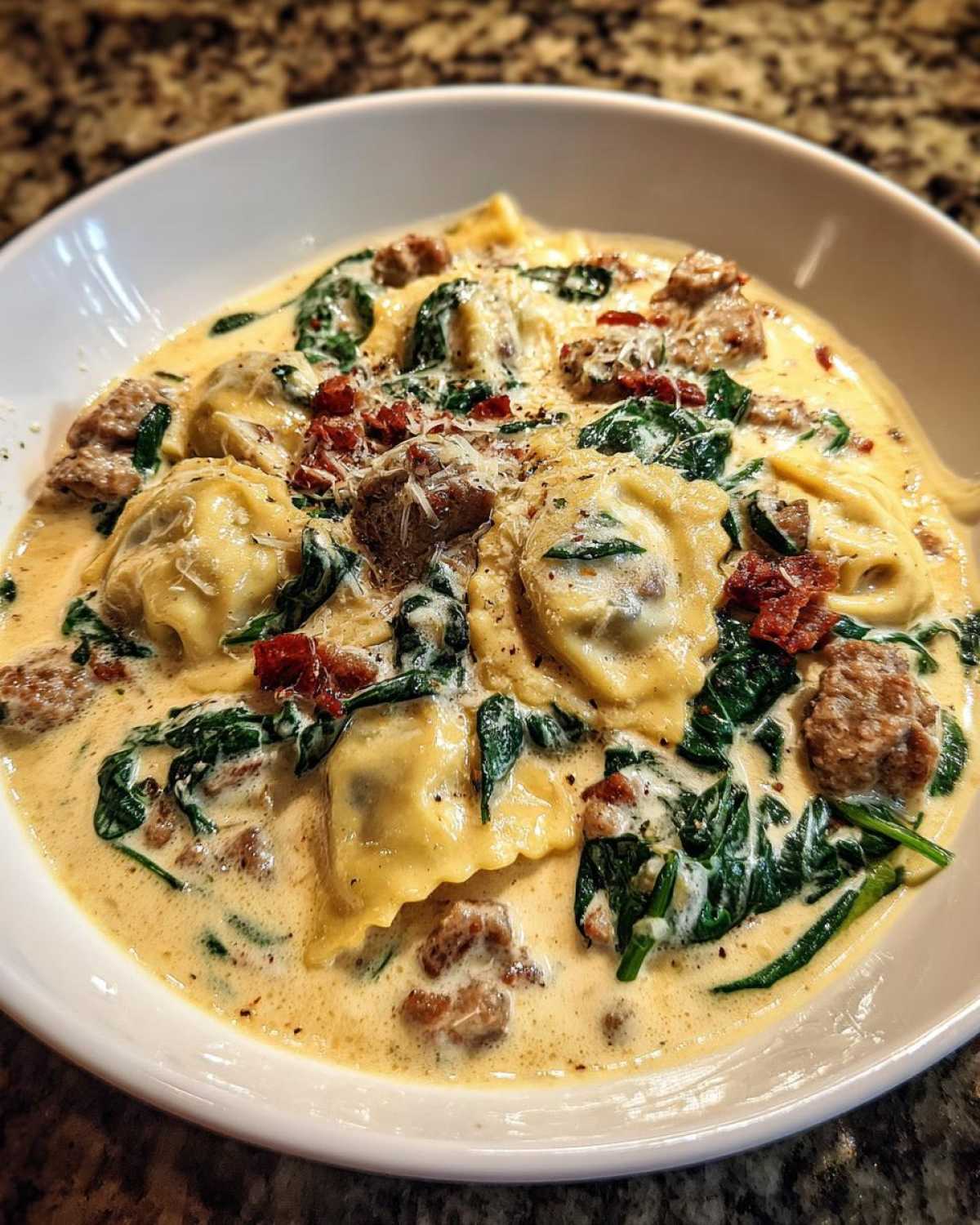 A close-up of a bowl of creamy ravioli soup featuring sausage, wilted spinach, and sun-dried tomatoes.