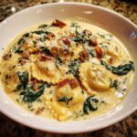 Close-up of a white bowl filled with rich ravioli soup, featuring cheese ravioli, spinach, sausage, and grated parmesan.