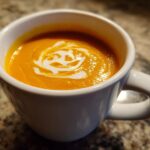 Close-up of vibrant orange squash soup topped with a swirl of cream in a white mug.