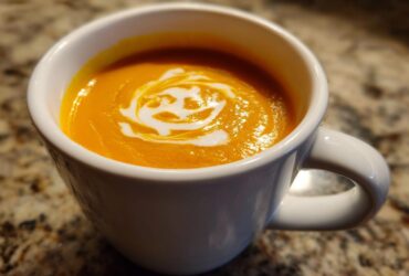 Close-up of vibrant orange squash soup topped with a swirl of cream in a white mug.