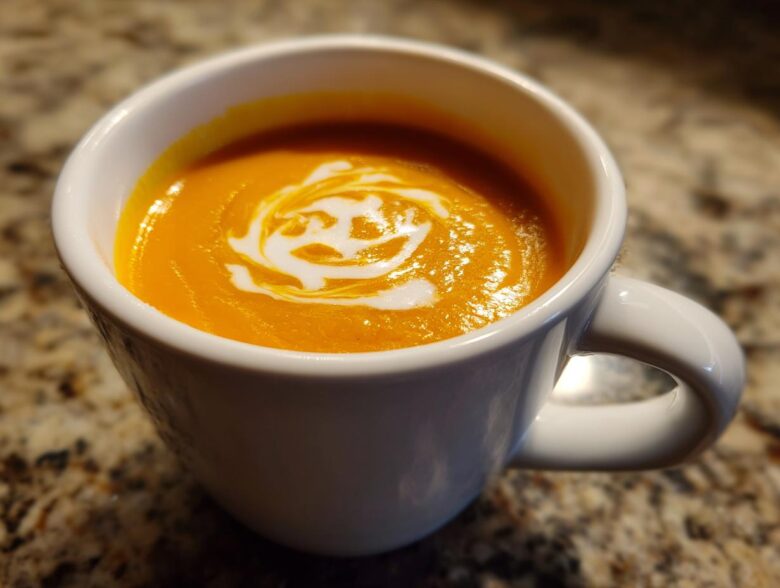 Close-up of vibrant orange squash soup topped with a swirl of cream in a white mug.